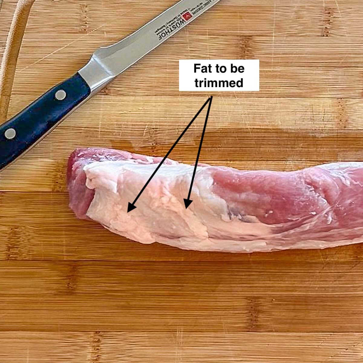 Raw pork tenderloin on a cutting board, with arrows showing the fat to be trimmed.