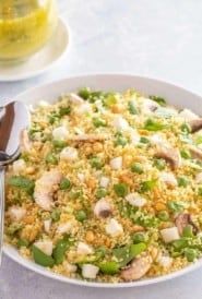 white bowl holding vegetable couscous salad, with 2 spoons on the side and dill vinaigrette in the back.