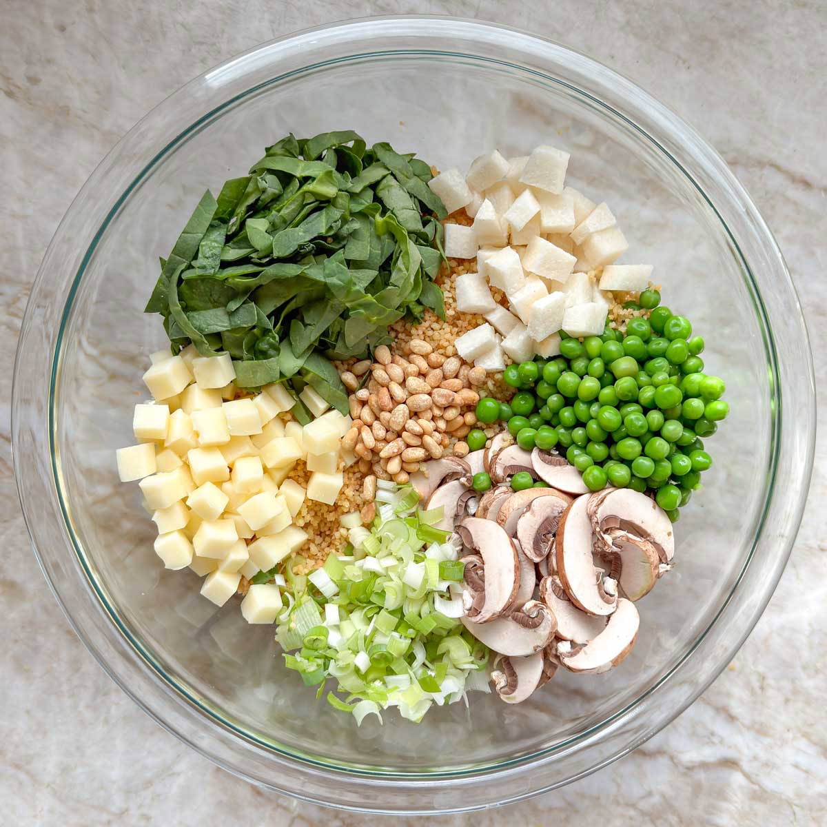 Toasted couscous in a mixing bowl with spinach, mushrooms, peas, jicama, green onions, mozzarella, and pine nuts.