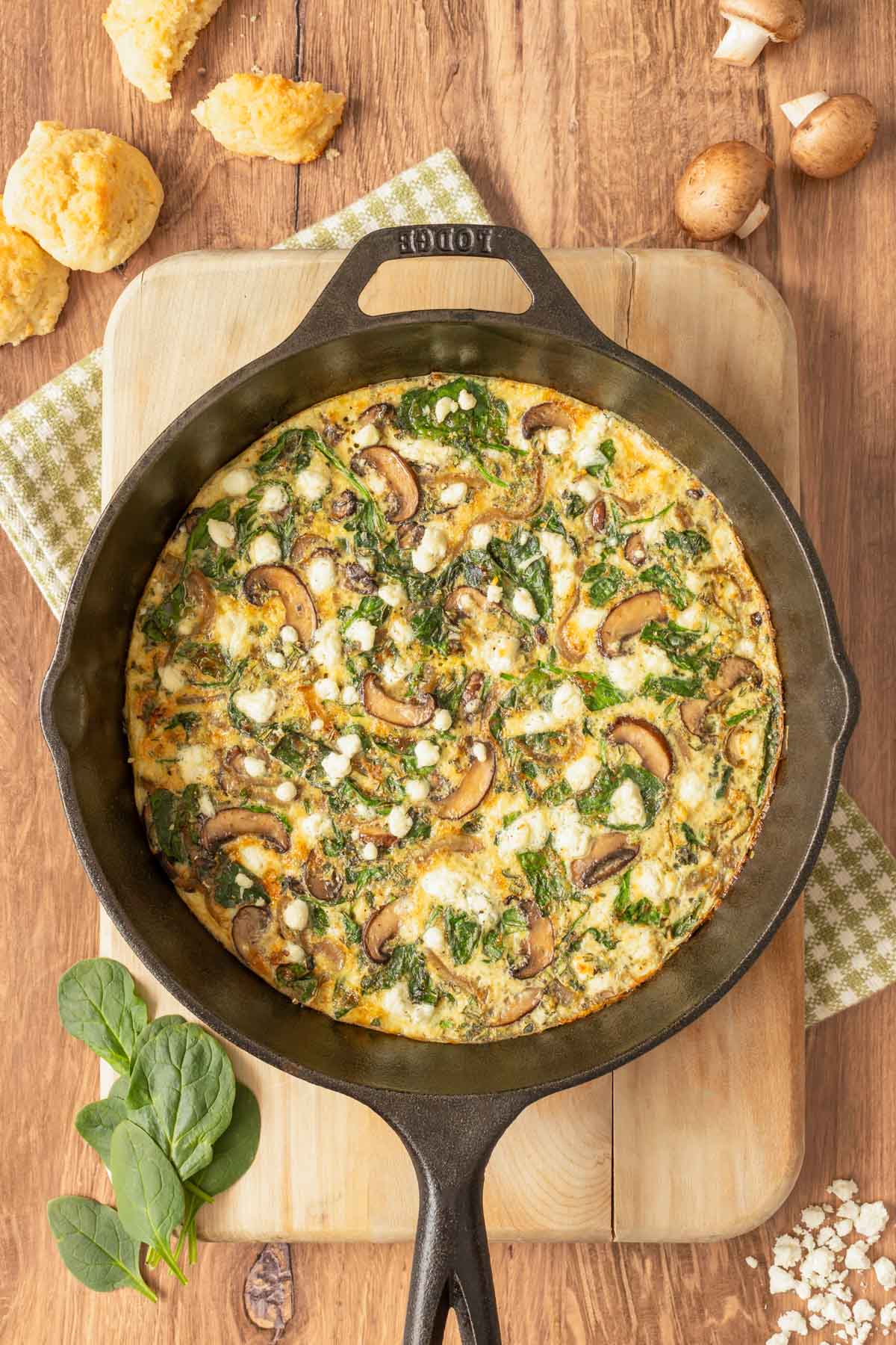 Overhead view of Vegetable Frittata in cast iron skillet set on a cutting board, with mushrooms, cheese, spinach and biscuits around the side