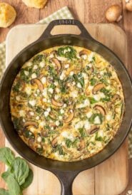 Overhead view of Vegetable Frittata in cast iron skillet set on a cutting board, with mushrooms, cheese, spinach and biscuits around the side