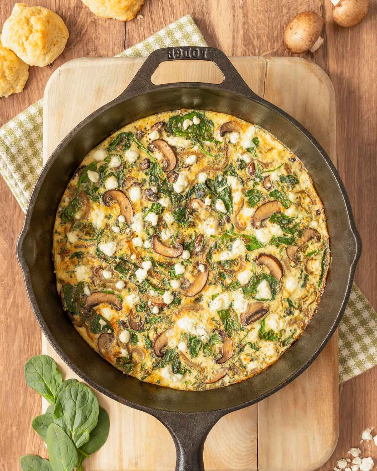 Overhead view of Vegetable Frittata in cast iron skillet set on a cutting board, with mushrooms, cheese, spinach and biscuits around the side